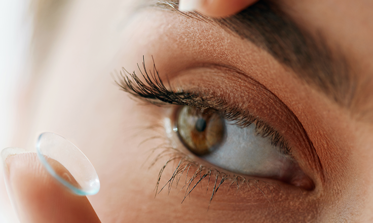 Close-up of a person’s eye as they prepare to insert a contact lens, with one finger gently holding the eyelid open and the contact lens resting on their fingertip