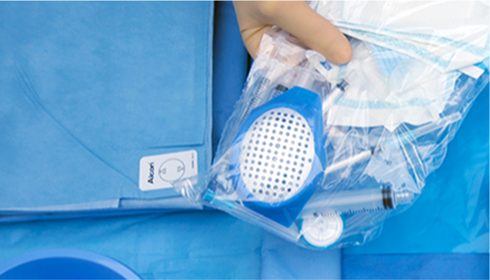 Hand holding a sterile surgical pack containing Alcon instruments and supplies, placed on a blue surgical drape