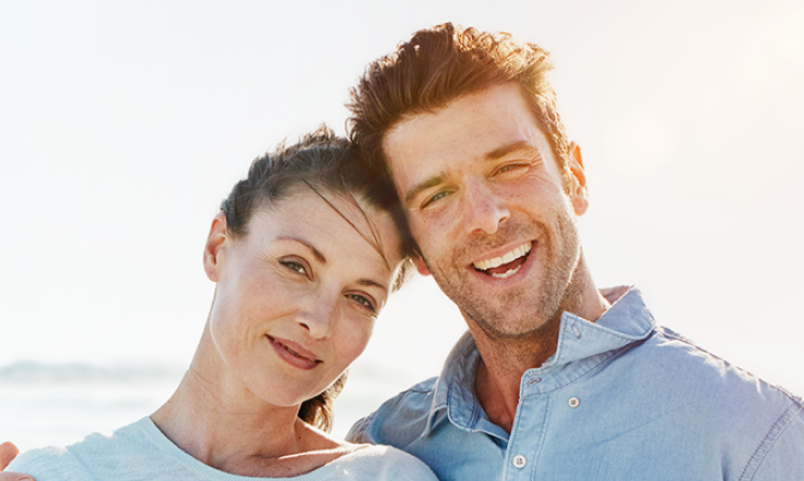Smiling couple standing close together outdoors on a sunny day, with the ocean in the background.