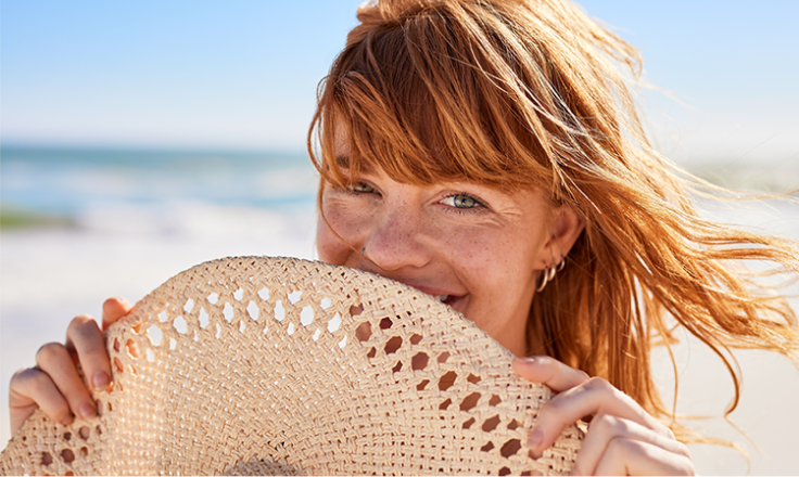 Smiling woman with red hair holding a woven sun hat in front of her face on a sunny beach