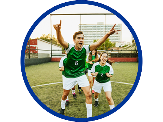 Excited soccer players in green jerseys celebrating on an outdoor field, with one player raising both arms in victory and teammates smiling behind him