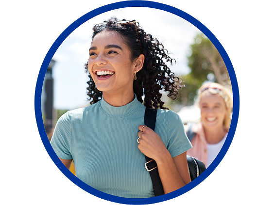 Smiling young woman with curly hair wearing a blue top and backpack, walking outdoors on a sunny day with a friend in the background.
