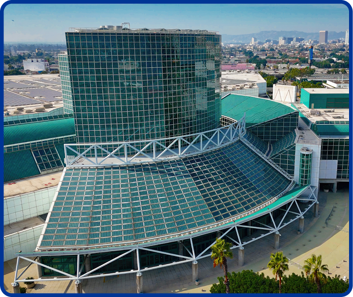 Aerial view of the Los Angeles Convention Center, featuring its distinctive green-tinted glass architecture and curved glass atrium. The city skyline is visible in the background under a clear sky.