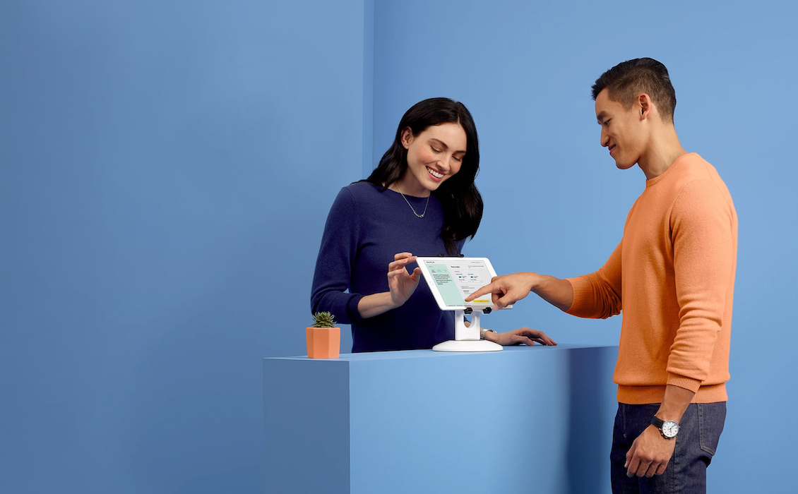 A smiling woman in a navy blue sweater stands behind a modern blue counter, assisting a man in an orange sweater as he interacts with a tablet device navigating the Meet Marlo interface