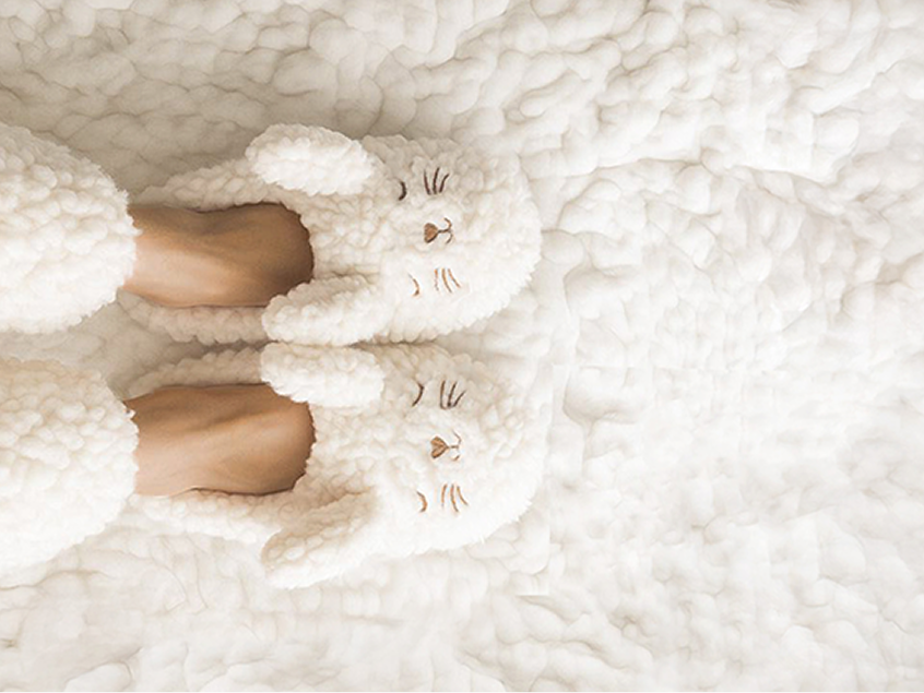 Person wearing fluffy white slippers shaped like sleepy animal faces with ears, resting on a soft, textured white blanket