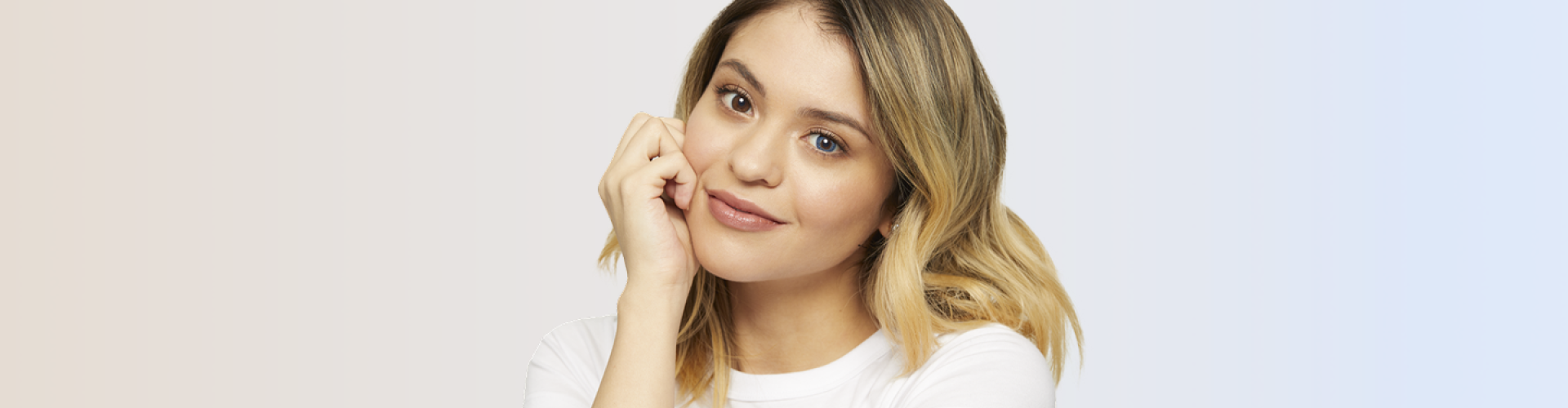 Smiling woman with blonde hair resting her chin on her hand, wearing a white top and one blue and one brown colored contact lens to show a side-by-side comparison effect.