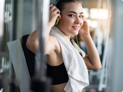 A young woman in a black sports bra sits on a workout machine at the gym, smiling while adjusting her headphones. A white towel is draped around her neck, and she appears to be taking a break from exercise. The background features gym equipment with sunlight streaming through the windows.
