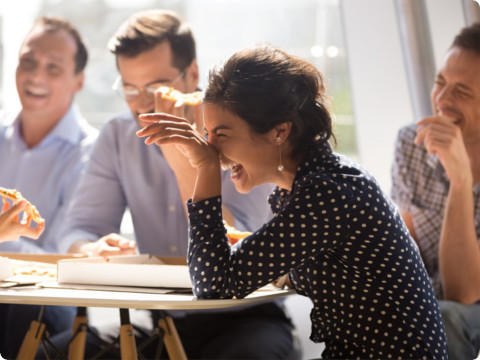 A group of colleagues shares a lighthearted moment while eating pizza in a bright office space. A woman in the foreground, wearing a polka-dotted blouse, laughs while holding a slice of pizza. The others, dressed in business casual attire, smile and engage in conversation around a table.