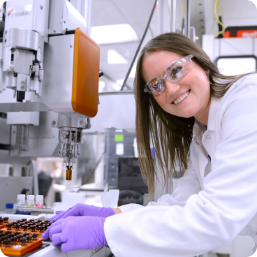 Eine Wissenschaftlerin mit weißem Laborkittel, Schutzbrille und lila Handschuhen lächelt bei der Arbeit in einem Labor. Sie hantiert mit kleinen Fläschchen unter einer Robotermaschine, umgeben von Laborgeräten in einer Hightech-Umgebung