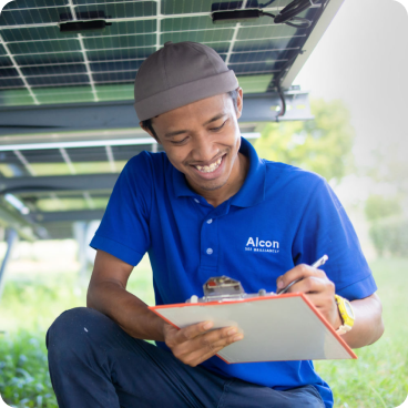 Ein Techniker in einem blauen Poloshirt der Marke Alcon und einer braunen Mütze lächelt, während er auf einem Klemmbrett schreibt. Er hockt im Freien unter einer Solaranlage, umgeben von Grünpflanzen