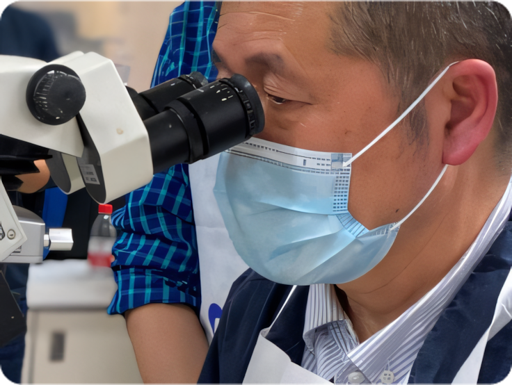 眼科トレーニングや研究セッションで、手術用マスクを着けた男性が顕微鏡を覗く。背後で観察する他の人々。