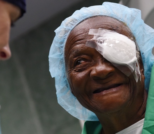 Close-up of an older person wearing a light blue surgical cap and an eye patch. The person is smiling gently.