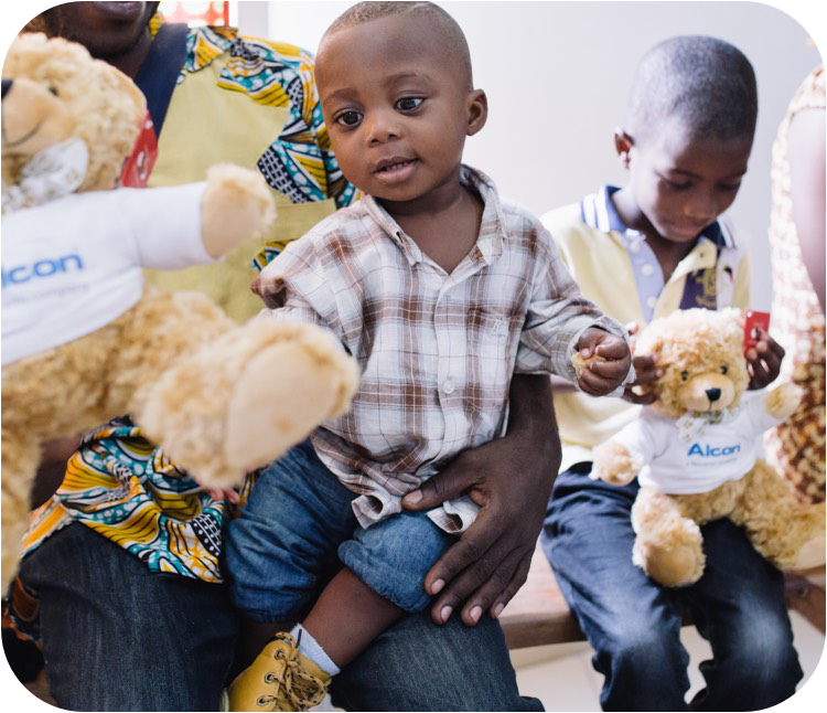 Jeune enfant assis sur les genoux d'un adulte, vêtu d'une chemise à carreaux et d'un jean retroussé, tendant la main vers un ours en peluche portant le logo Alcon sur sa chemise. Un autre enfant à l'arrière-plan, tenant également un ours en peluche similaire, est assis et regarde l'ours.