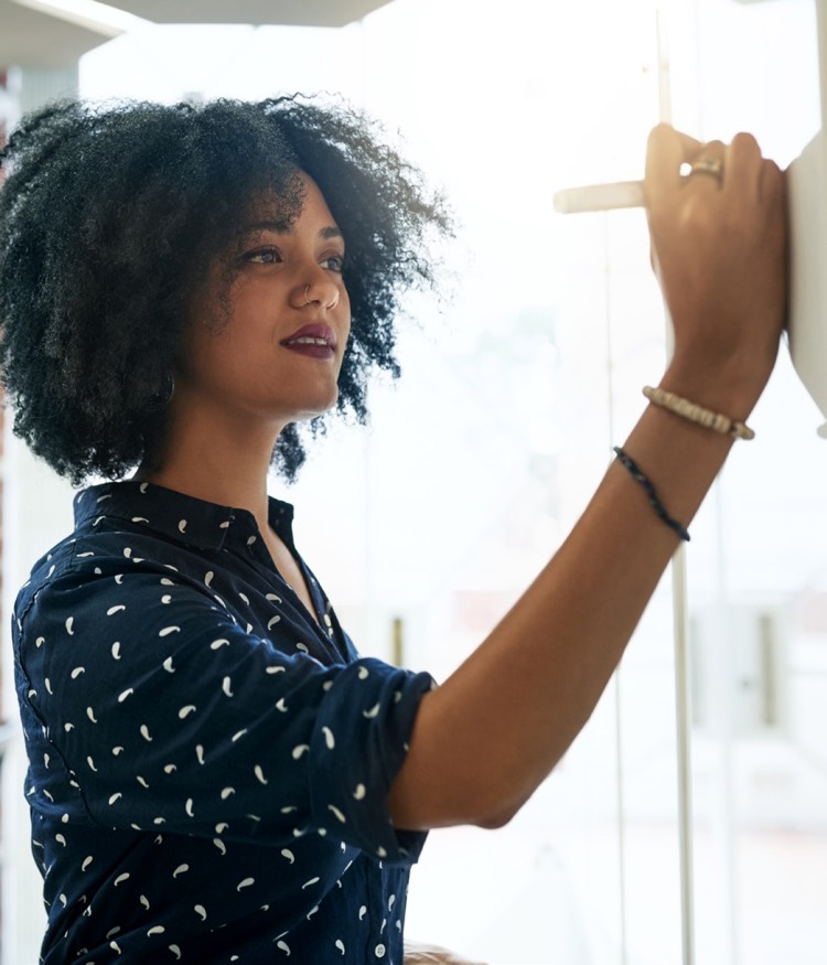 A woman with curly hair and a patterned navy blue blouse writes or draws on a whiteboard in a well-lit space.