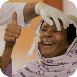 Une femme âgée joyeuse portant un foulard à fleurs sourit tandis qu'un professionnel de la santé, portant des gants, examine délicatement son œil. Elle lève le pouce, exprimant ainsi son bonheur et sa gratitude. Un mouchoir en papier est glissé dans son foulard, signe d'une intervention médicale récente.