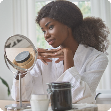 woman with long dark hair in ponytail in white robe looking into round vanity mirror