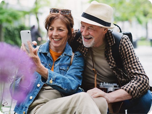 Older couple sitting together outdoors, leaning close while taking a selfie with a smartphone; greenery and flowers in the background.
