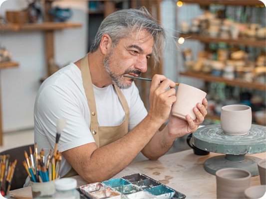 Middle-aged man with gray hair wearing an apron painting a ceramic pot at a pottery workstation with brushes and tools.