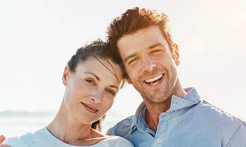 Smiling couple standing close together outdoors on a sunny day, with the ocean in the background.