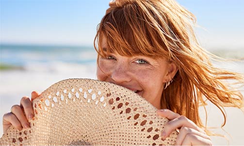Smiling woman with red hair holding a woven sun hat in front of her face on a sunny beach