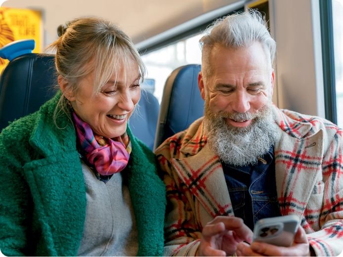 Smiling older couple sitting together on a train, looking at a smartphone and laughing