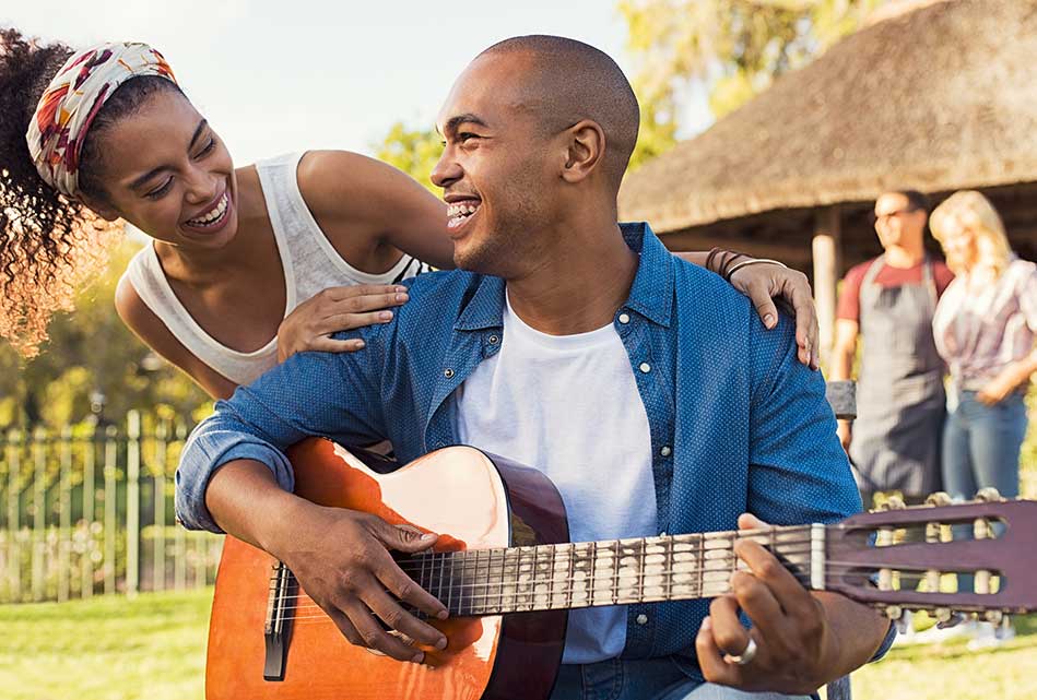 Man sitting and playing guitar as a woman smiles and rests a hand on his shoulder at an outdoor gathering