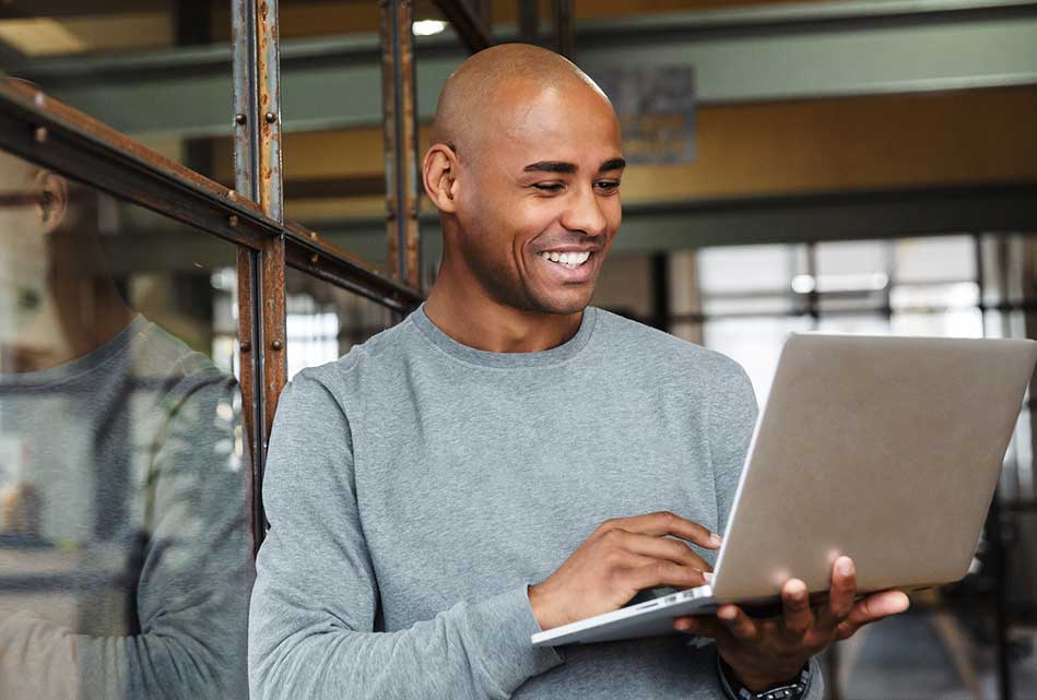 A man standing indoors, smiling while typing on a laptop near large windows.