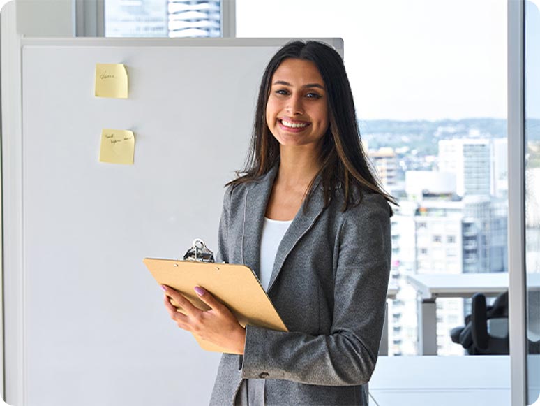 A woman standing in an office environment, smiling and holding a clipboard with sticky notes on a whiteboard behind her.